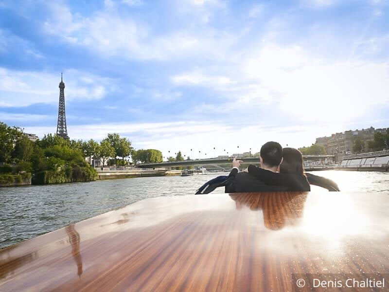 Un couple est assis à l'avant d'un bateau-mouche et tourne le dos à l'objectif. Ils regardent et pointent du doigt la tour Eiffel.